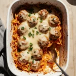 Cheesy baked spaghetti and meatballs in a white dish with fresh parsley on a wooden table.