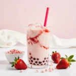 Pale pink strawberry milk tea in a clear glass with ice, pink straw, fresh strawberries, and tapioca pearls.